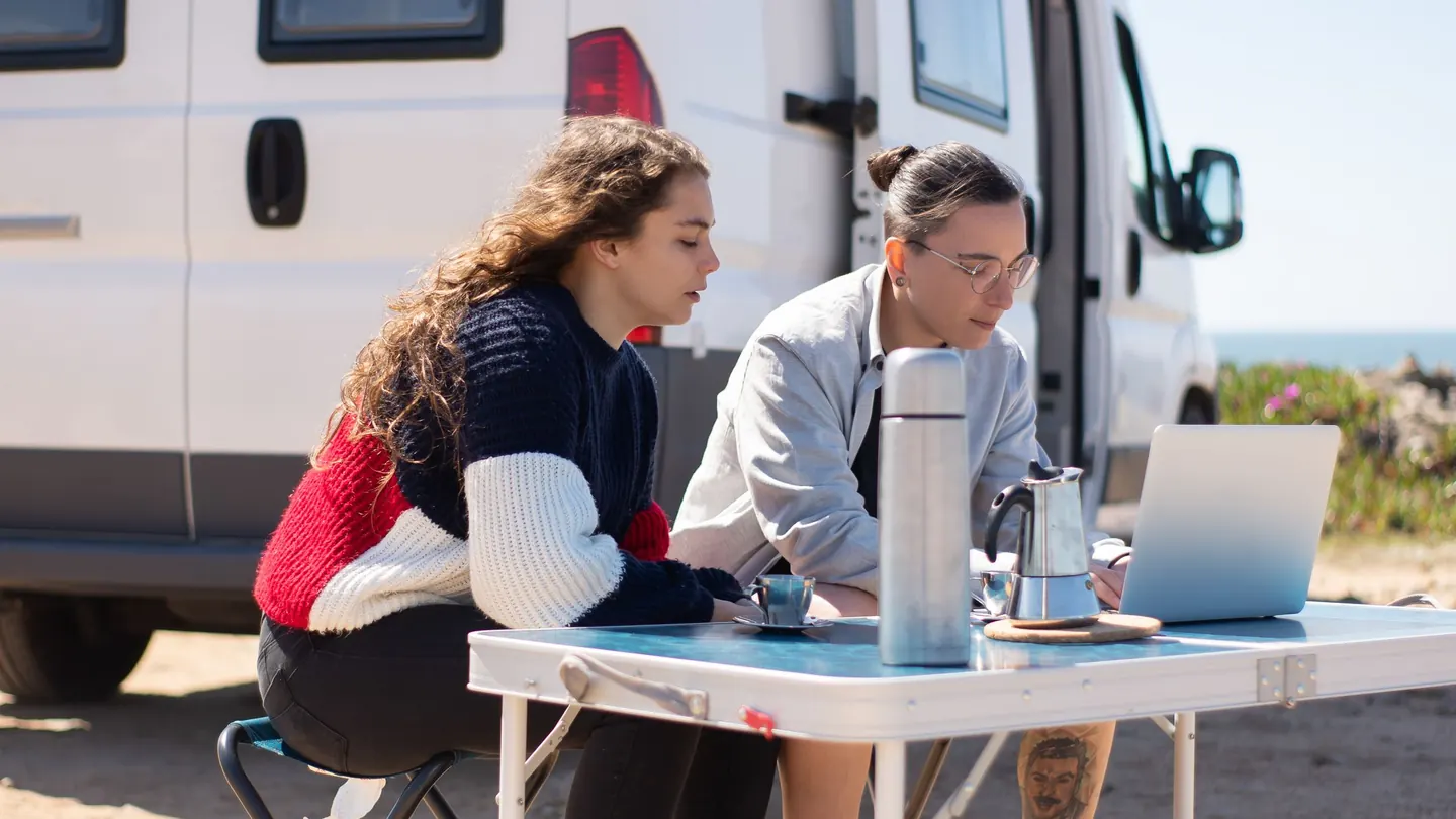 Two travelers use a laptop at an outdoor table beside a campervan.